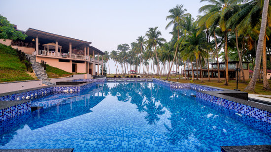 A resort with a blue-tiled swimming pool surrounded by palm trees and a multi-storied building with balconies.