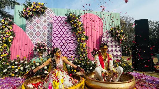 alt-text a bride and groom sitting on big bowls during haldi ritual with rose petals placed around them - heritage village resorts & spa, Manesar