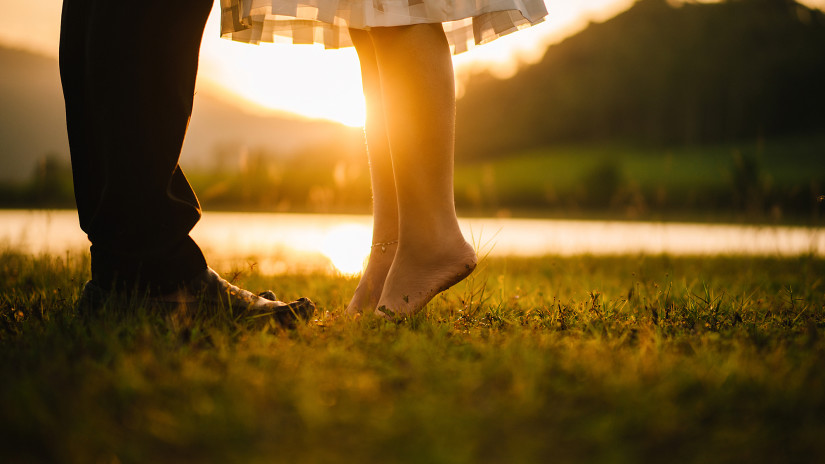 Close-up of two people's lower legs at sunset, one barefoot on tiptoe.