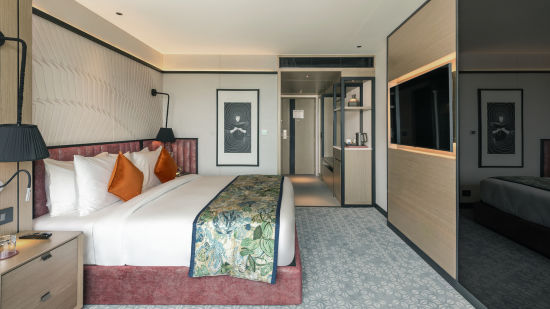 alt-text Side view of a Deluxe room ,featuring a comfortable bed, a side table with a lamp, an LCD TV, modern amenities, a well-equipped and decorated space, and a mirror