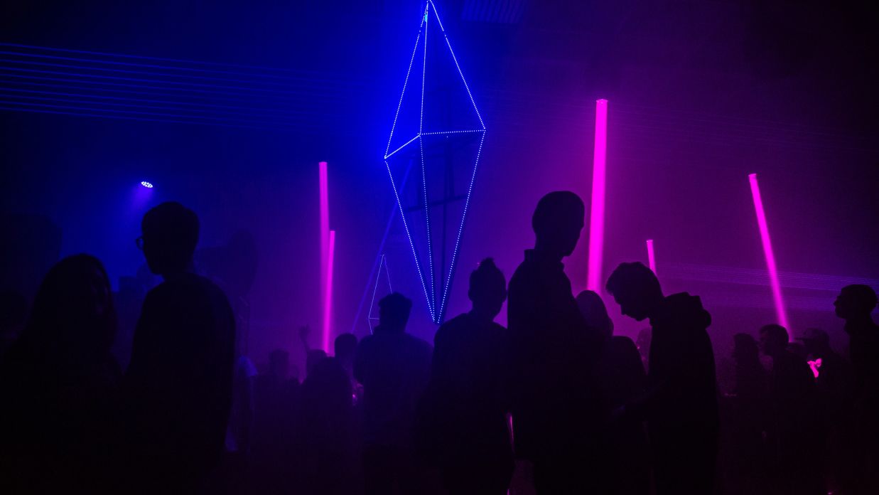 Interior view of a club featuring silhouettes of people and a triangular bipyramid shaped neon light placed in the background tiirsv