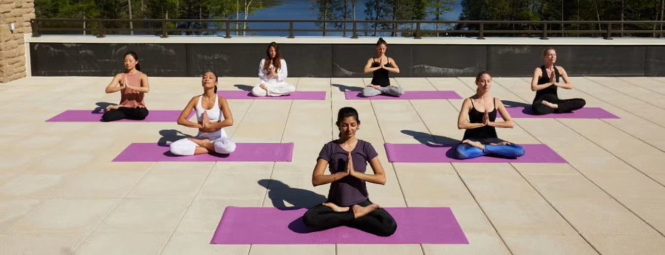 A group of people seated on yoga mats in various meditation poses on an outdoor terrace with a view of a lake and forest.