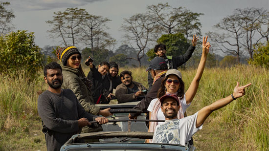 A group of joyful people on a jeep safari, waving and smiling, surrounded by grasslands with a cloudy sky above.