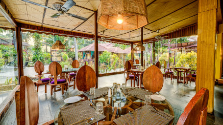 An open dining area in a resort with wooden furniture, ceiling fans, and a view of lush tropical greenery outside.