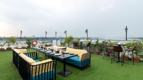  An outdoor dining setup on a rooftop garden with a grassy floor, overlooking a bridge and river during daylight.