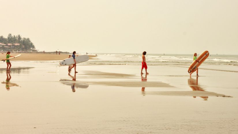 four people walking on the beach holding surfing boards with the evening sky in the background - Water Sports Activities in Goa