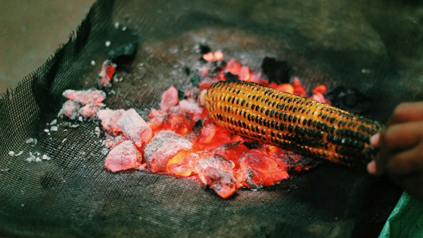 a corn on the cob being grilled directly on hot coal by a street vendor