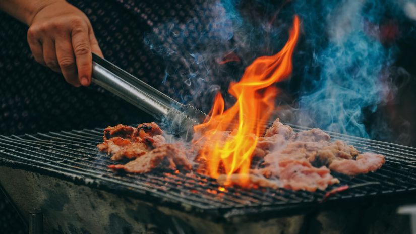 An action shot of meat grilling over an open flame with visible smoke and a person's hand holding tongs.