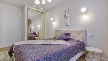 Side view of a double bed with purple duvet in a room with white walls and bright lights