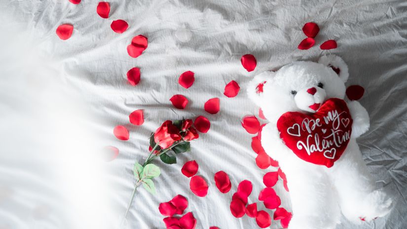 a teddy bear holding a be my valentine plush heart on a bed strewn with rose petals