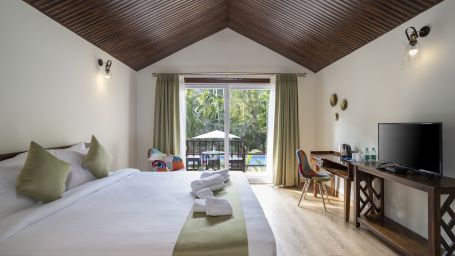 A view of our room with a large bed with green runners placed in front of a television, hanging lights to illuminate the room and a sliding door opening to the balcony - Stone Wood Nature Resort, Gokarna