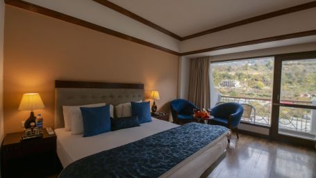 A room featuring a clean bed, side table lamps, sofa chairs, table and a balcony - Mastiff Grand, Manali