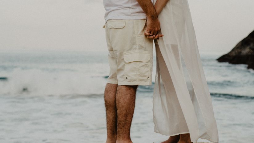 a couple holding hands wearing white clothes on a beach while the ocean can be spotted in the background and captured from a lower angle