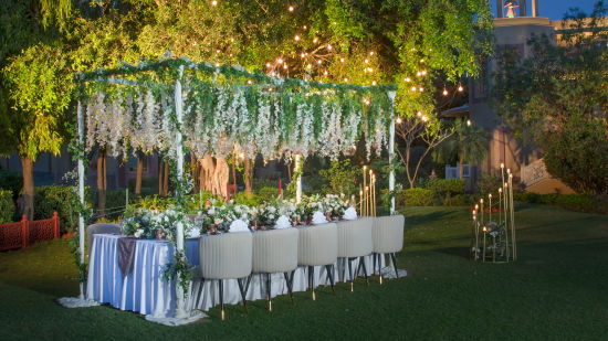 alt-text An outdoor evening event with a long dining table set for a meal, adorned in a white tablecloth, surrounded by nature and illuminated by overhead string lights.