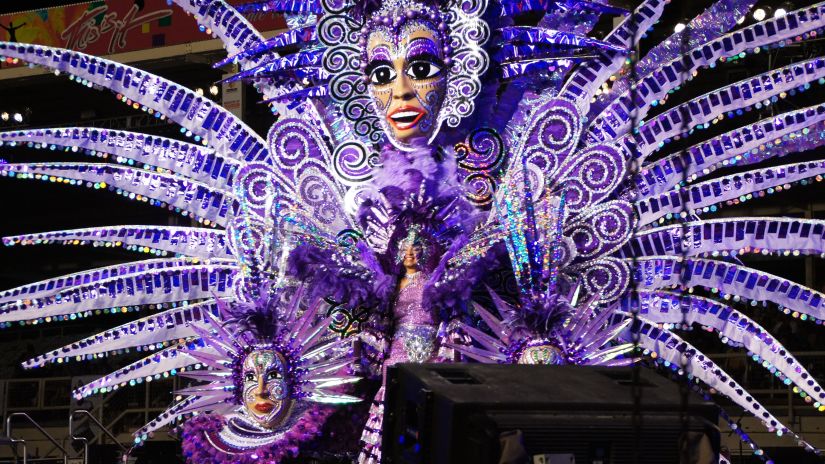 corner view of a carnival decoration in purple colour with large face masks with design on them and a woman dancing on the centre of the stage