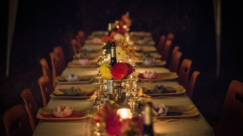 A long tabel with chairs on either side featuring cutlery and a bottle of wine