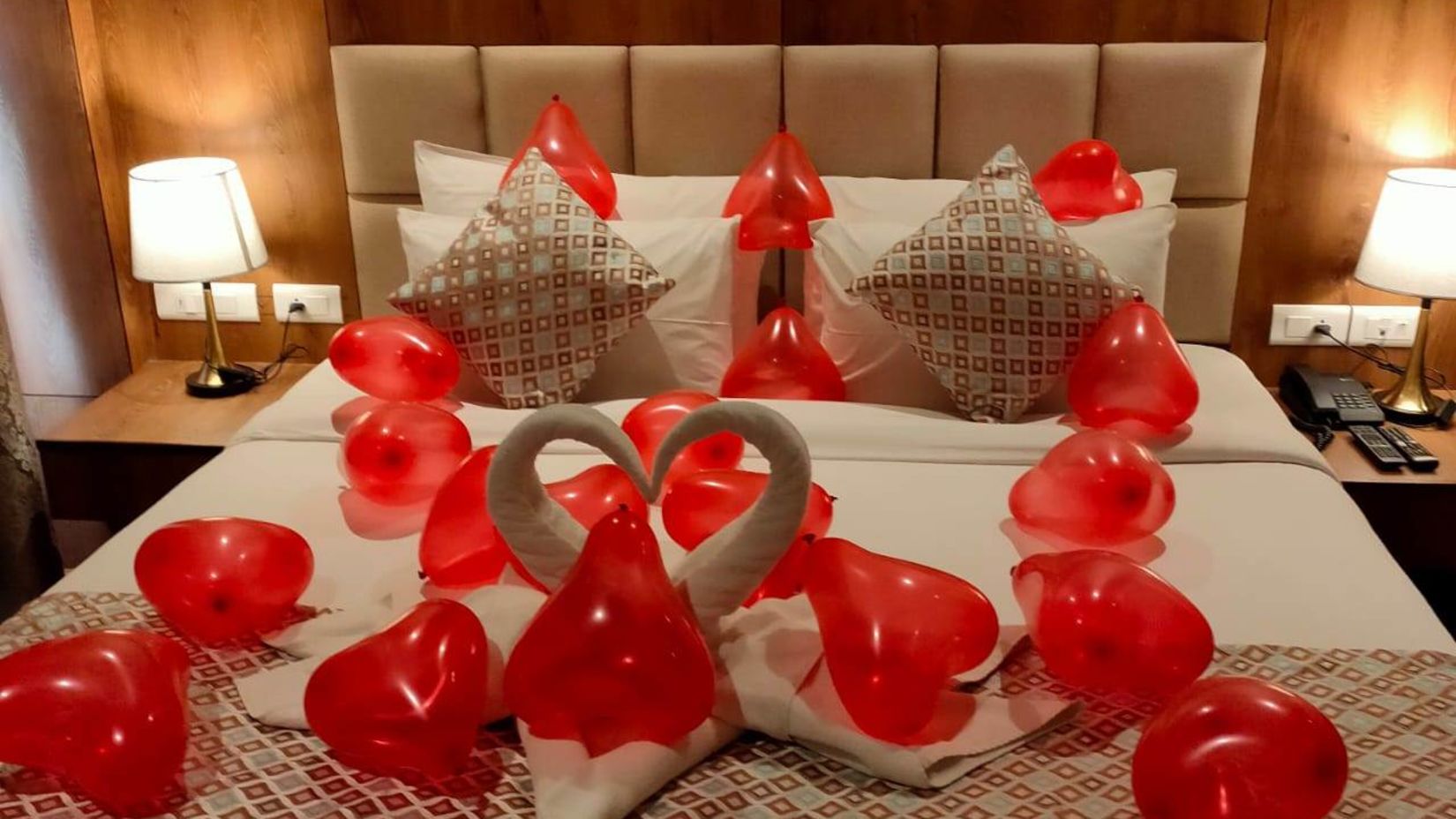 A room decorated with red heart shaped balloons with a swan placed in the middle of the bed - The Orchid Hotel, Manali