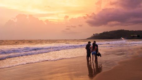 a view of baga beach with 3 people standing near the shore with sunset in the background