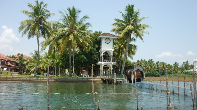 A tower with a clock next to a vembanad lake with coconut trees next to it