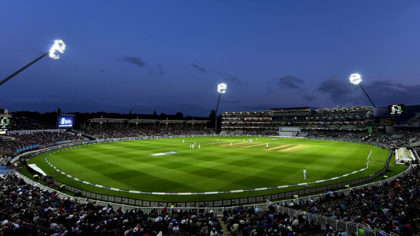 a far out view of a cricket stadium with floodlights on and dark skies in the background