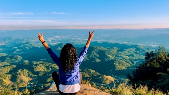 a girl sitting on top of a hill looking at the nature below and the blue sky in the background