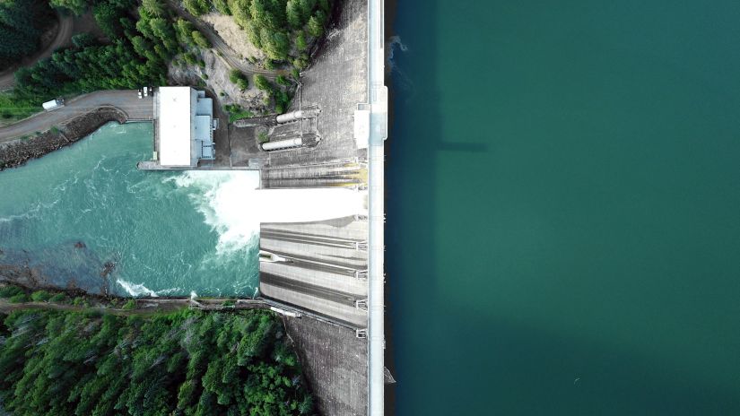 view from a dam from above with water cascading down to a waterbody with trees on either side below