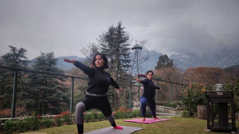 an image of two women doing yoga on a yoga mat in the morning with snow-capped mountains in the background covered with clouds @ Lamrin Norwood Green, Palampur