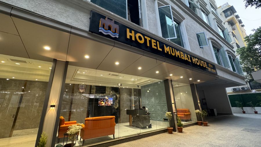 The entrance of Hotel Mumbai House with a modern design, showcasing a sign above the door, glass walls, a sofa set in the foyer, and potted plants on the sidewalk.