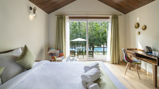 A hotel room with a large bed, a work desk and a chair and a pool side view - Stone Wood Nature Resort, Gokarna