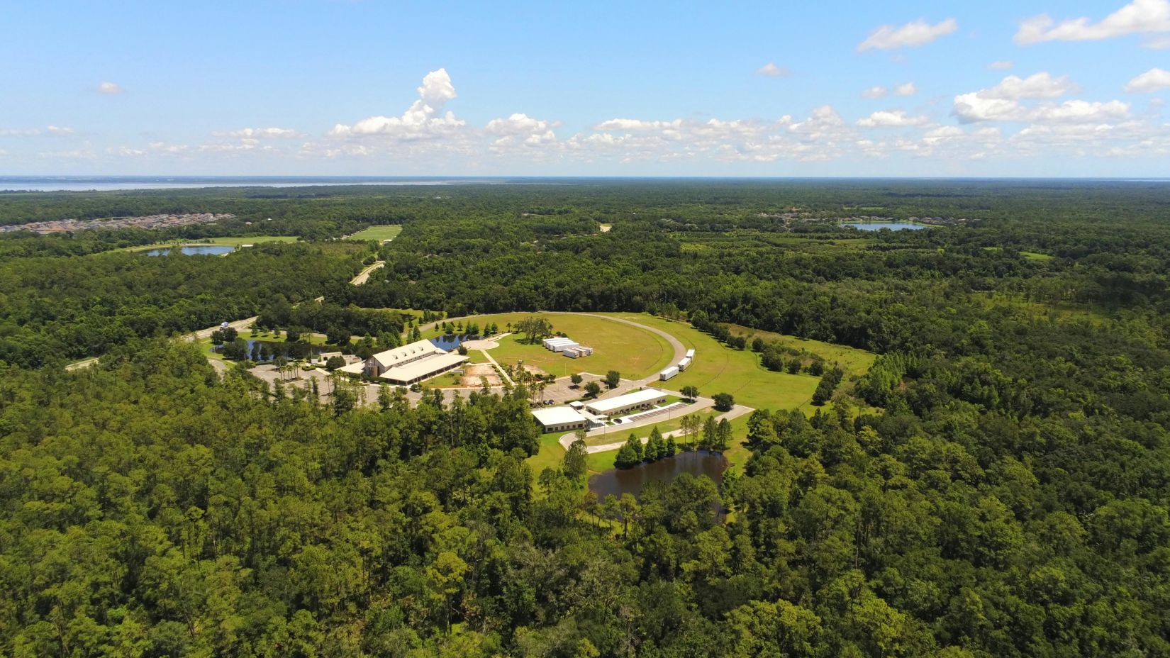 An aerial shot of a facility ringed by a road amidst expansive greenery and distant lakes.