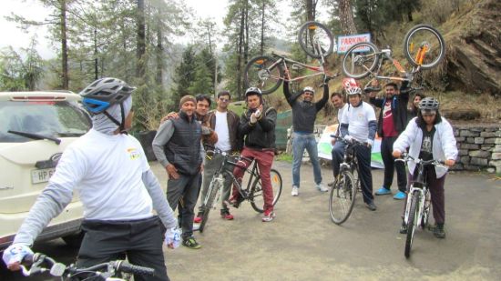 A lively group of cyclists celebrates a pause in their mountain ride, displaying their bikes and enjoying the camaraderie on a forested roadside.