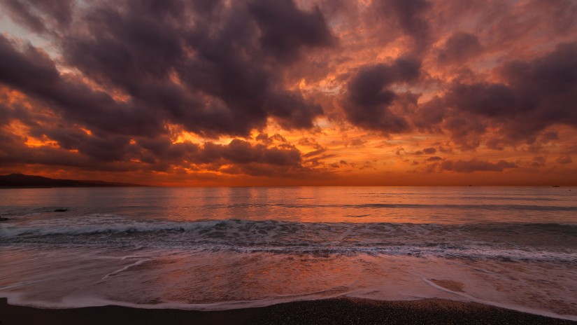 A wide view of a beach with a backdrop of cloudy evening sky and a peachy sunset