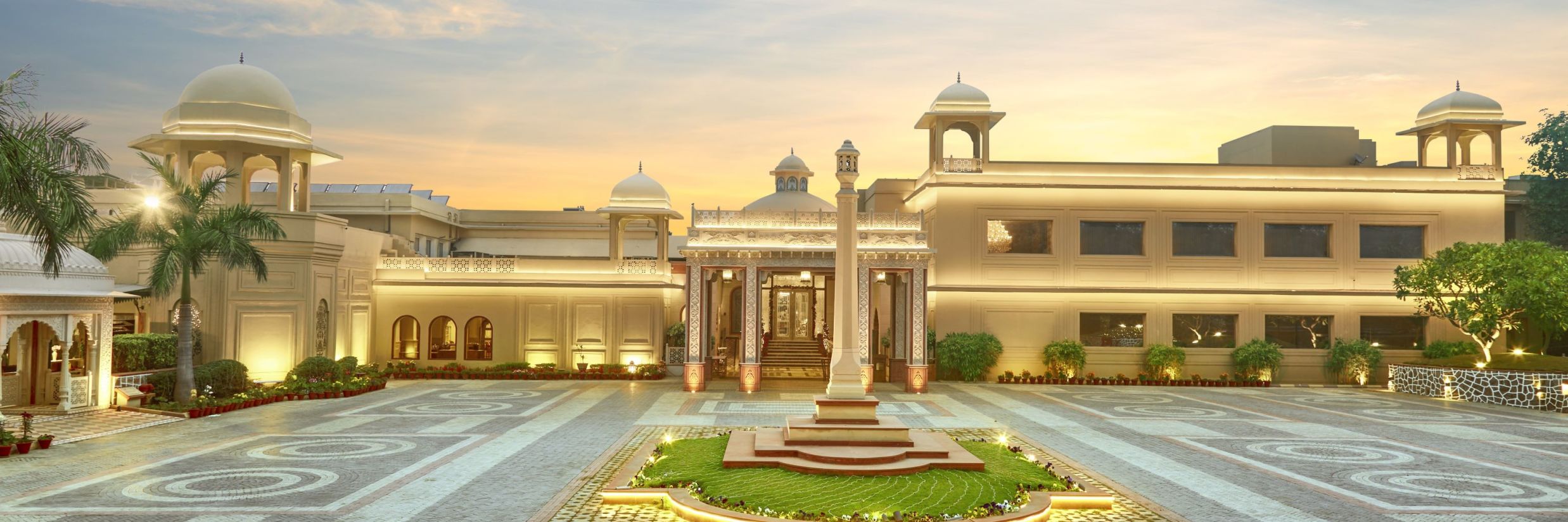 Manesar - Front view of a grand hotel building at dusk, featuring a symmetrical design with a central fountain, landscaped gardens, and ornate light posts leading to the main entrance, all under a soft, glowing sky.