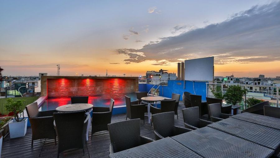 A rooftop terrace during sunset with wicker furniture, a lit pool and a view of the city skyline - TGI Apple Inn, Jaipur 