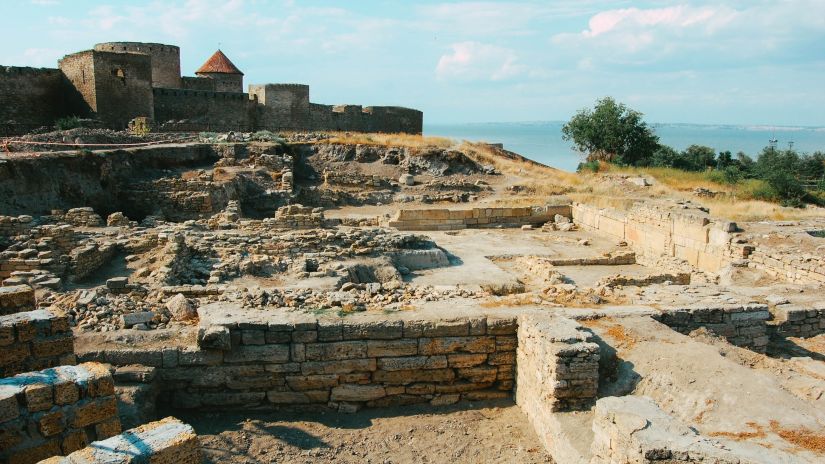 Ancient stone ruins under a blue sky, remnants of a historical site with partial walls and structures on a vast archaeological excavation site.