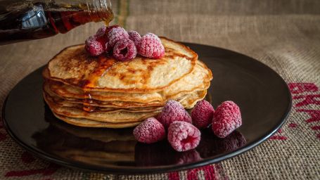 syrup being poured on a stack of pancakes topped with raspberries