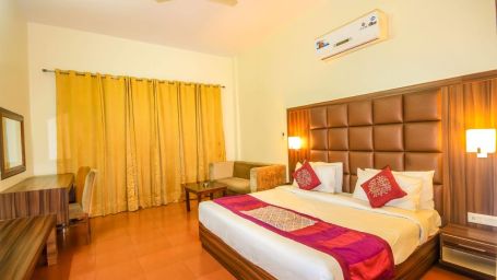 A beautiful deluxe room with a wooden bed yellow curtains table lamps a writing desk a sofa and a mirror - Little Mastiff, Dharamshala