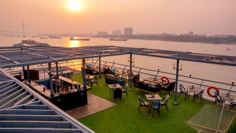 A bar counter on a rooftop terrace during sunset, showcasing a selection of alcohol bottles and a river in the background.