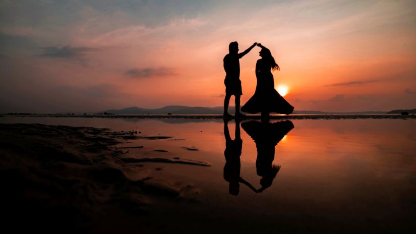 a couple dancing on a beach with the sun setting in the background and pastel hues in the sky