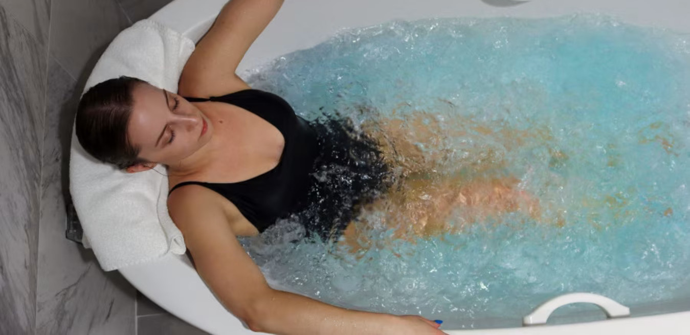 Woman in a black swimsuit enjoying a bubbling hydrotherapy tub, leaning back with eyes closed, against a marble tile background.