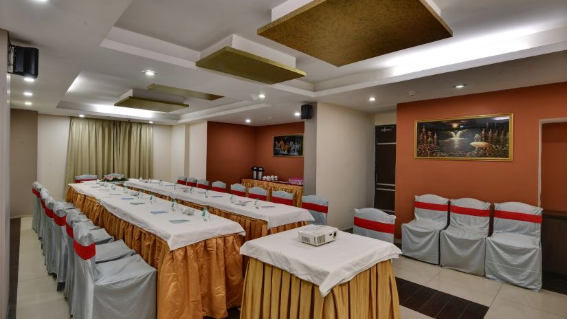 A conference room with a U-shaped table setup and a projector in the foreground, with grey chair covers and red sashes - Click hotels Yuvraj, Surat