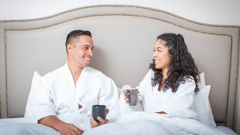 a couple sitting together on a bed under the blanket with coffee mugs in hand