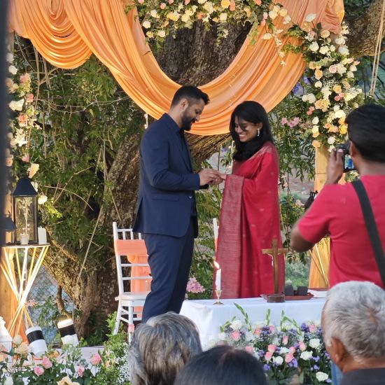 alt-text Bride and groom exchanging rings at an altar under a tree, surrounded by seated guests in an outdoor wedding in Coonoor.