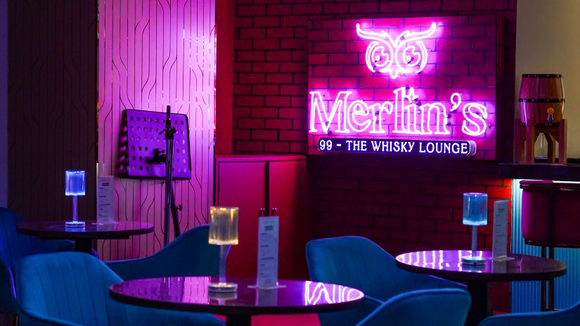 blue chairs and wooden table placed on a marbled floor with merlin's 99 signboard glowing in pink in the background