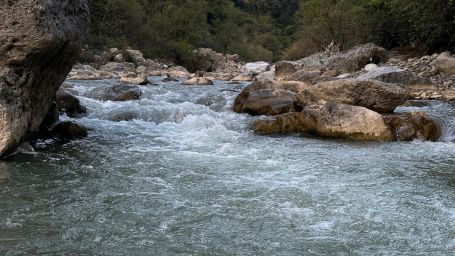 A swift, clear mountain stream flows over and around large rocks in a forested area with hills in the background.