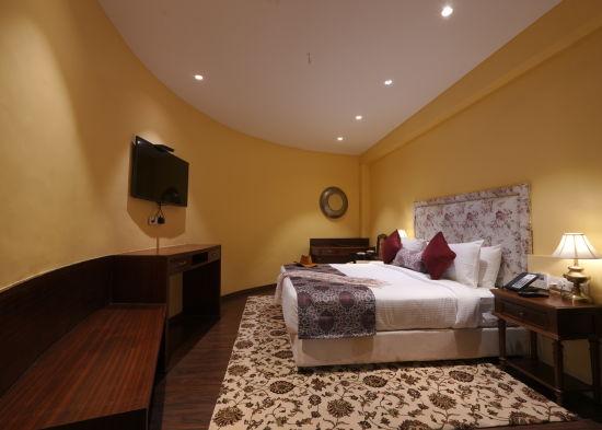 alt-text Side view of the double bed, a wall mounted television and a wide table in the Suite Cottage Room at The Garden Ananta Elite Rajkot 4
