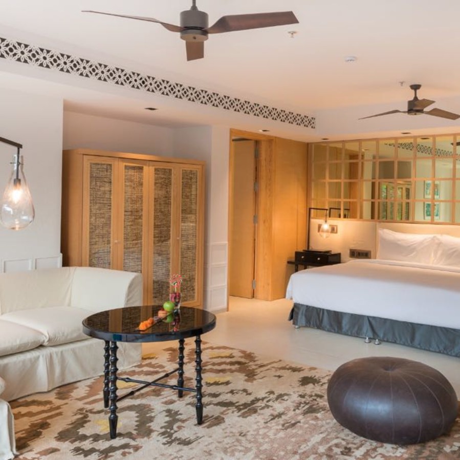 alt-text Room with a brown pouffe and black round table surrounded by a white couch and bed - Azaya Beach Resort Goa