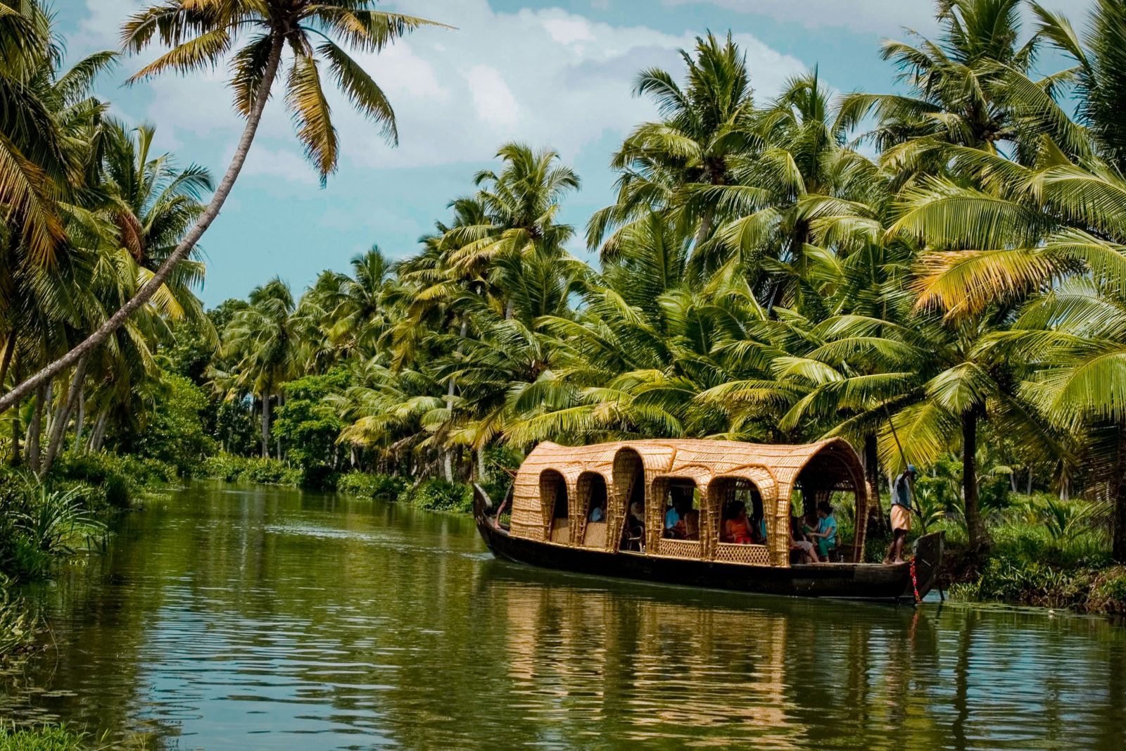 A house boat containing people floating on a narrow stream flanked by coconut trees on either sides