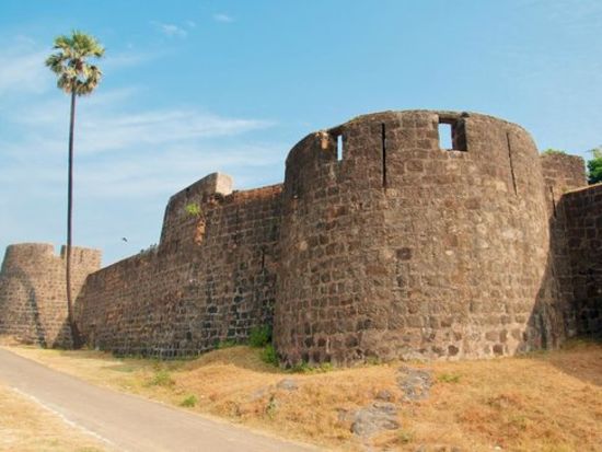 alt-text an far out view of Madh Fort with a small road on the side and blue sky in the background