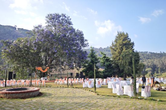 alt-text  Scenic view of an outdoor wedding venue during the day with guests, chairs, and an altar under a blooming purple-flowered tree in Coonoor.